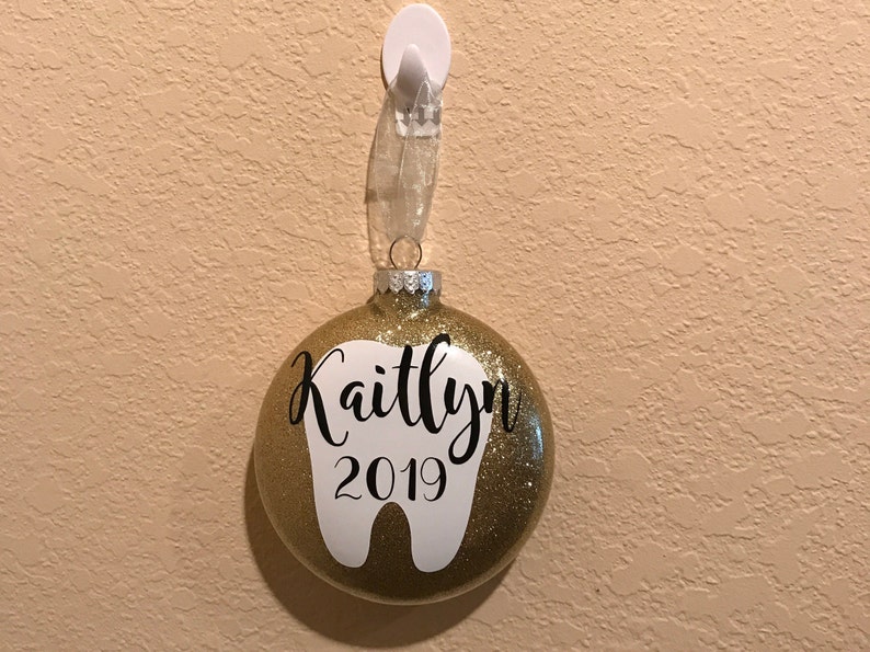 Puede incluir: Adorno navide&ntilde;o de purpurina dorada con forma de diente blanco y el nombre Kaitlyn y el a&ntilde;o 2019.