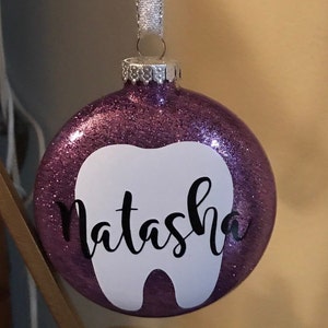Puede incluir: Adorno de Navidad de purpurina morada con forma de diente blanco y el nombre "Natasha" escrito en cursiva negra.