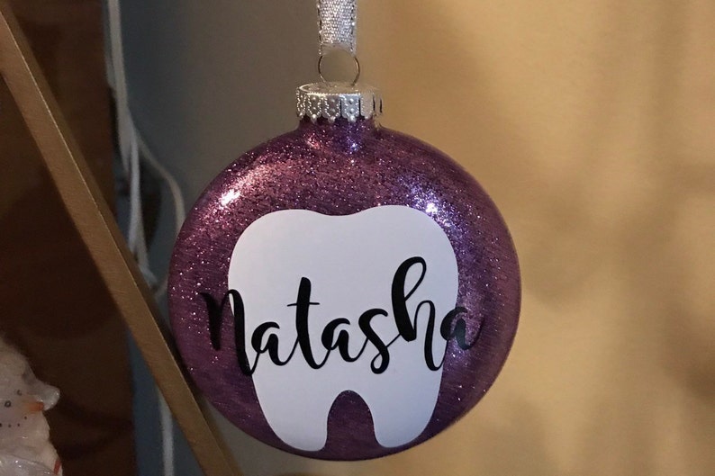 Puede incluir: Adorno de Navidad de purpurina morada con forma de diente blanco y el nombre "Natasha" escrito en cursiva negra.