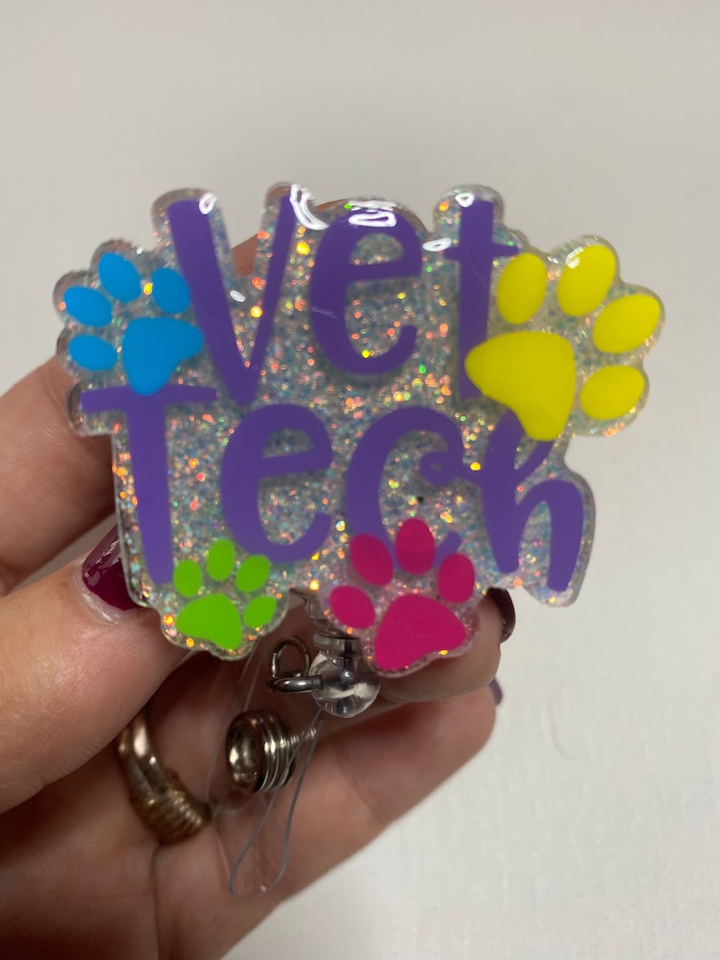 K&ouml;nnte beinhalten: Ein lila glitzernder Badgehalter mit dem Text "Vet Tech" und Pfotenabdr&uuml;cken in Blau, Gelb, Gr&uuml;n und Pink.