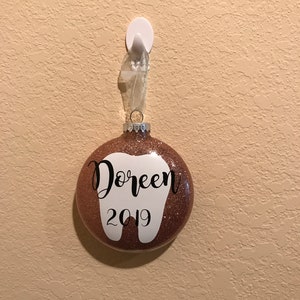 Puede incluir: Un adorno navide&ntilde;o de purpurina dorada rosa en forma de diente con el nombre "Doreen" y el a&ntilde;o "2019" escrito en &eacute;l.