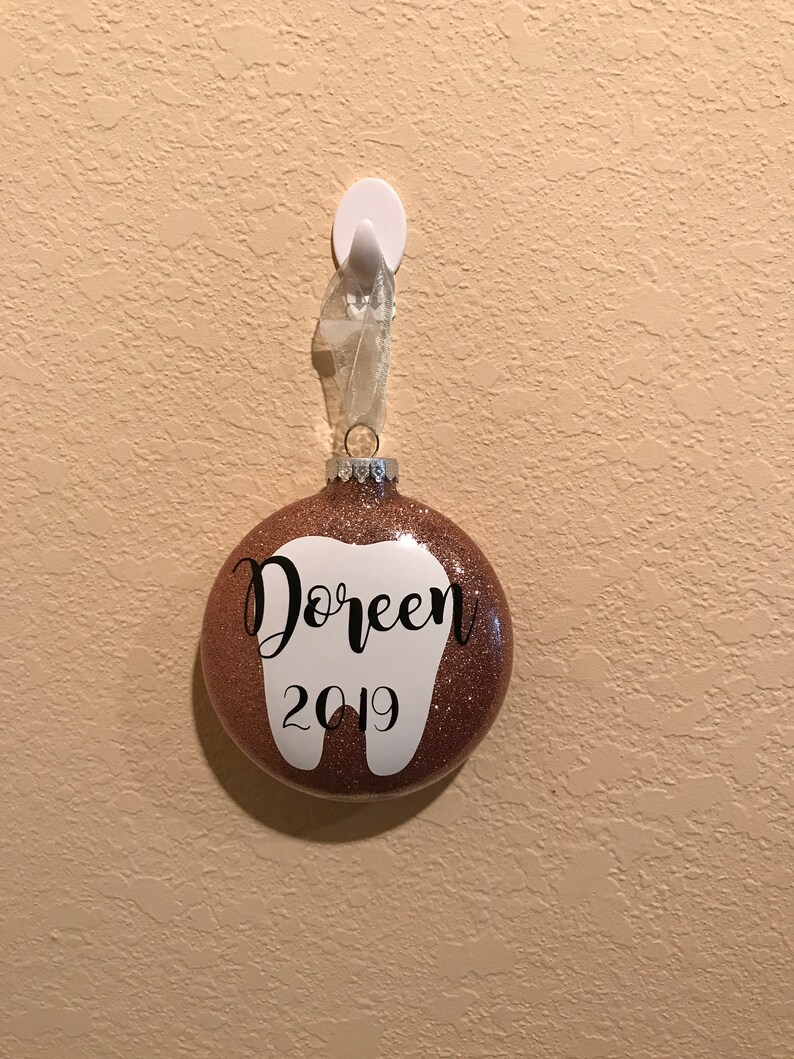 Puede incluir: Un adorno navide&ntilde;o de purpurina dorada rosa en forma de diente con el nombre "Doreen" y el a&ntilde;o "2019" escrito en &eacute;l.