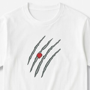 Puede incluir: Camiseta blanca con un diseño gráfico de garras grises y un corazón rojo. Las garras están en ángulo y parecen rasgar la tela. La camiseta tiene cuello redondo y mangas cortas.