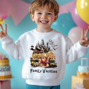 May include: White sweatshirt featuring a colorful graphic of a castle, characters, and the text "UNIVERSAL STUDIOS" and "Family Vacation". The design includes Minions and other animated figures. The sweatshirt is worn by a smiling child.