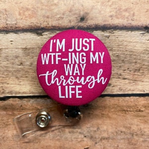 May include: A circular hot pink badge reel with a fabric-covered front featuring the white text I'M JUST WTF-ING MY WAY through life. The badge reel includes a clear plastic strap with a metal snap button and a retractable cord mechanism.