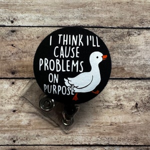May include: A black and white round button with a white duck and the text "I think I'll cause problems on purpose". The button is attached to a clear plastic retractable badge holder.