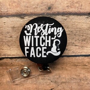May include: A round black badge reel featuring white text that reads Resting WITCH FACE. The design includes small white silhouettes of two bats and a witch hat with a dangling spider. A clear plastic strap with a metal snap button is attached below.