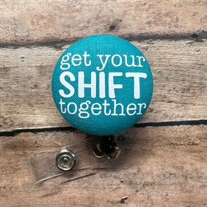 May include: Teal blue fabric button with white text that reads "get your SHIFT together". The button is attached to a clear plastic retractable badge holder with a black cord.