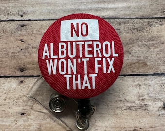 May include: A circular red badge reel with a fabric-covered top featuring the white text NO ALBUTEROL WON'T FIX THAT. The device includes a clear plastic strap with a metal snap button and a retractable cord mechanism for holding identification cards.