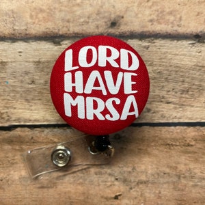 May include: Red button badge with white text that reads "LORD HAVE MRSA". The badge is attached to a retractable badge holder.