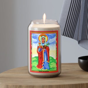 May include: A lit, clear glass candle with a painted image of a saint. The image features a woman in red and blue robes, holding a chalice and a cross. The candle sits on a wooden surface.