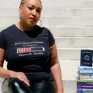 Może przedstawiać: Kobieta w czarnym t-shircie z napisem "Ed.D Doctor of Education in Progress Future Dr. Buckley" i grafiką paska postępu. Siedzi na schodach z książkami i paszportem na stopniach obok niej. Ma na sobie czarne skórzane spodnie i czarne szpilki.
