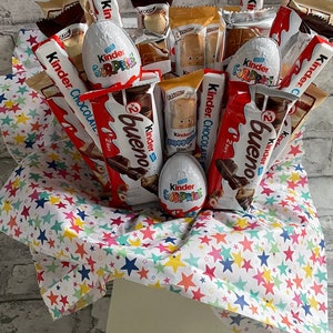 May include: A gift basket filled with various chocolate treats, including Kinder Surprise eggs, Kinder Bueno bars, and Kinder Chocolate bars. The basket is lined with white paper with colourful stars.