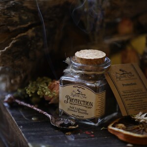 Puede incluir: Un frasco de vidrio con tapa de corcho lleno de incienso en polvo marr&oacute;n. El frasco tiene una etiqueta que dice "Protection Self Lighting Powder Incense" y una peque&ntilde;a ilustraci&oacute;n de un p&aacute;jaro. El frasco est&aacute; sentado sobre una superficie de madera con flores secas y una cuchara.