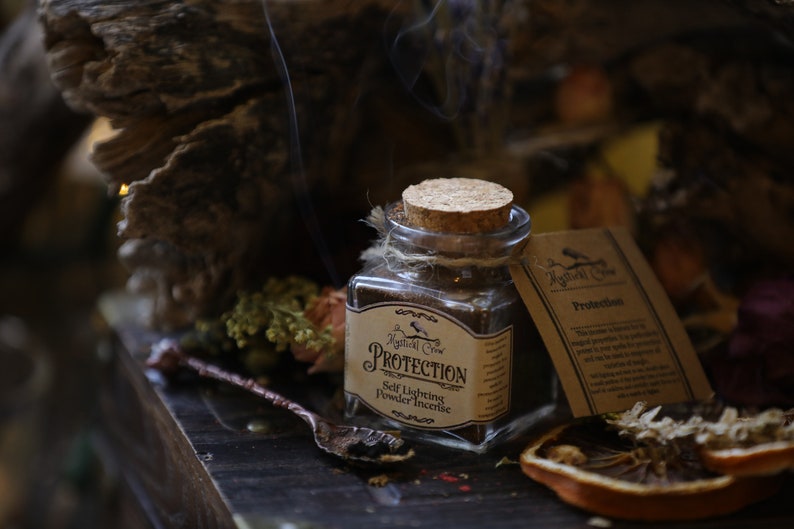 Puede incluir: Un frasco de vidrio con tapa de corcho lleno de incienso en polvo marr&oacute;n. El frasco tiene una etiqueta que dice "Protection Self Lighting Powder Incense" y una peque&ntilde;a ilustraci&oacute;n de un p&aacute;jaro. El frasco est&aacute; sentado sobre una superficie de madera con flores secas y una cuchara.