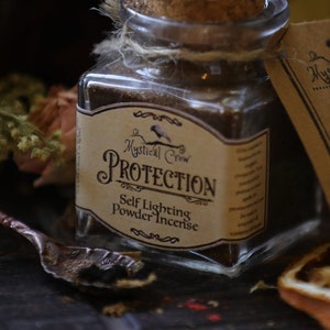 Puede incluir: Un frasco de vidrio lleno de incienso en polvo marr&oacute;n etiquetado como "Mystical Crow Protection Self Lighting Powder Incense". El frasco tiene una tapa de corcho y est&aacute; atado con una cuerda.