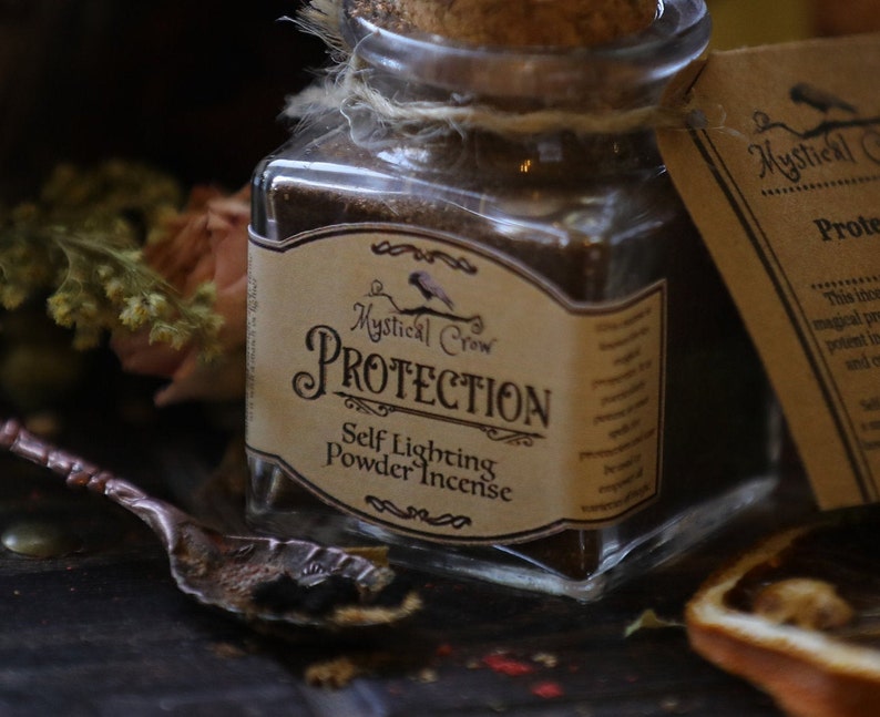 Puede incluir: Un frasco de vidrio lleno de incienso en polvo marr&oacute;n etiquetado como "Mystical Crow Protection Self Lighting Powder Incense". El frasco tiene una tapa de corcho y est&aacute; atado con una cuerda.