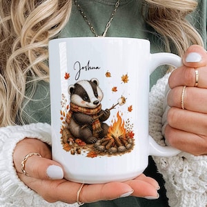 May include: A white ceramic mug featuring a graphic illustration of a badger wearing a plaid scarf, sitting by a campfire and roasting a marshmallow. The name Joshua is printed in black script above the badger, surrounded by orange autumn leaves.