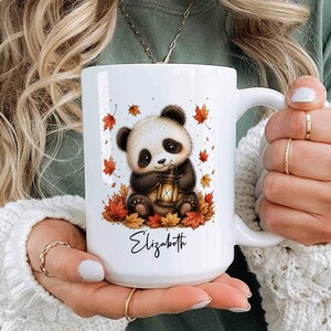 May include: A white ceramic mug featuring a graphic illustration of a cute panda bear sitting in a pile of colorful autumn leaves while holding a small lantern. The name Elizabeth is printed in black cursive script below the panda illustration.
