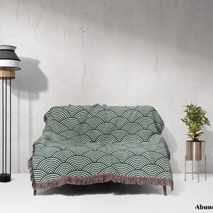 May include: A living room setting with a sofa covered by a green and white patterned throw. A black and white floor lamp is on the left, and a plant in a gold stand is on the right. The word "Abundance" is at the bottom of the image.