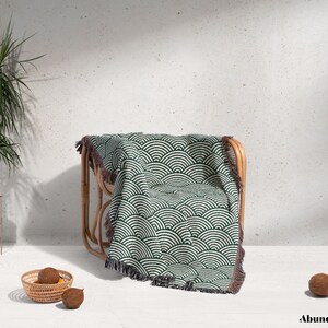 May include: A green and white patterned throw blanket with a fringed edge is draped over a natural rattan chair. The blanket features a repeating wave design. A potted plant and a small basket with fruit are in the foreground.