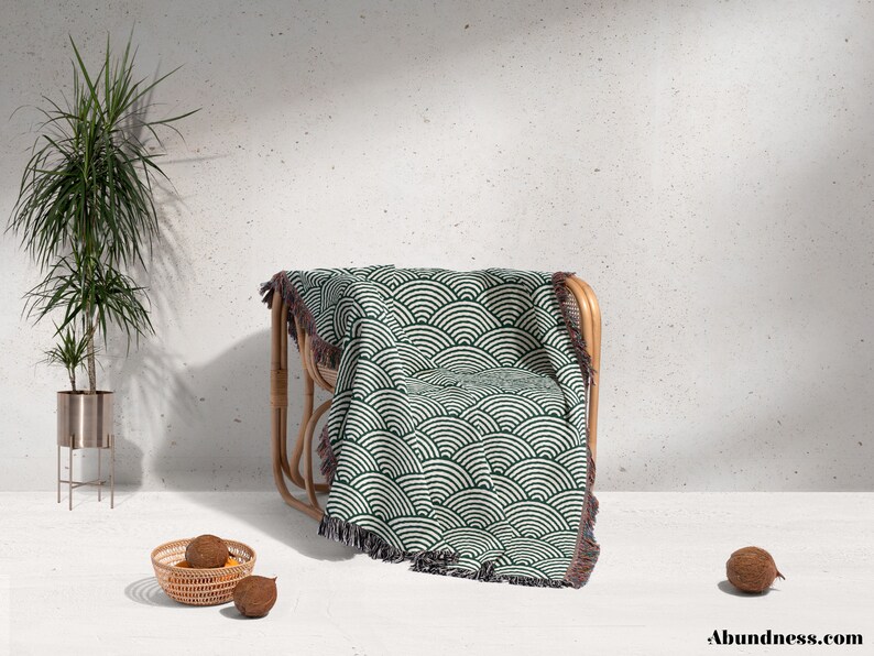 May include: A green and white patterned throw blanket with a fringed edge is draped over a natural rattan chair. The blanket features a repeating wave design. A potted plant and a small basket with fruit are in the foreground.