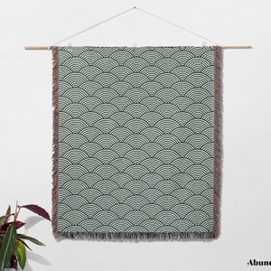 May include: A rectangular wall hanging with a repeating wave pattern in dark green and white. The edges have a fringed trim. The hanging is suspended from a wooden rod by a string.