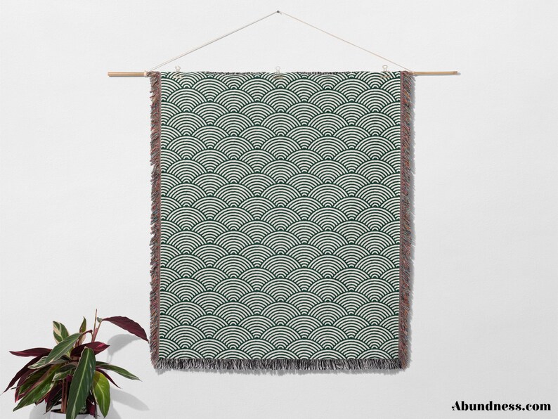May include: A rectangular wall hanging with a repeating wave pattern in dark green and white. The edges have a fringed trim. The hanging is suspended from a wooden rod by a string.