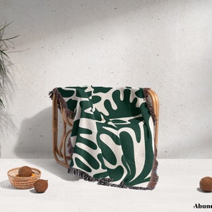 May include: A green and white woven throw blanket with an abstract pattern of leaves and branches. The blanket is draped over a wooden chair with a white background.