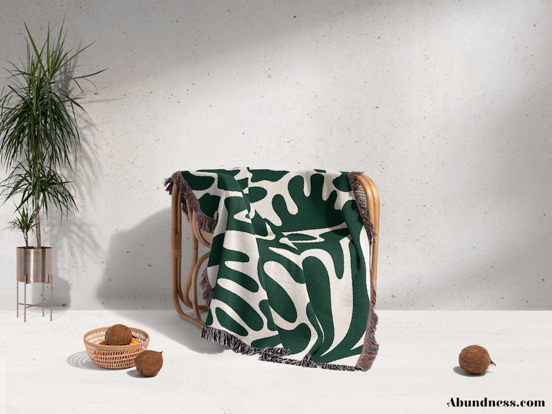May include: A green and white woven throw blanket with an abstract pattern of leaves and branches. The blanket is draped over a wooden chair with a white background.