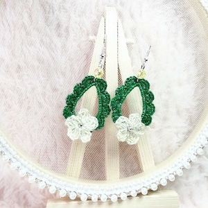 Puede incluir: Un par de pendientes de ganchillo verde con flores de ganchillo blancas. Los pendientes cuelgan de ganchos plateados.