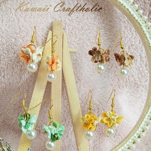 Puede incluir: Pendientes florales hechos a mano en tonos naranja, marrón, verde y amarillo, cada uno con una perla. Se exhiben en un pequeño caballete de madera y dentro de un aro decorativo con el texto "Kawaii Craftholic".