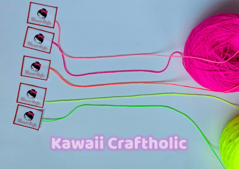 May include: Four spools of yarn in pink, orange, green, and yellow colors. The yarn is arranged in a line with the spools on the left and the yarn extending to the right. The yarn is labeled "Kawaii Craftholic".