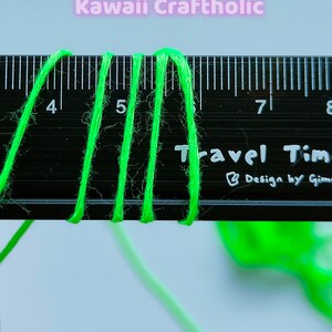 May include: Close-up of three strands of bright green yarn measured against a black ruler. The ruler has markings for inches and the numbers 4, 5, 7, and 8 are visible. The text "Travel Time" and "Design by Gimme" are printed on the ruler.