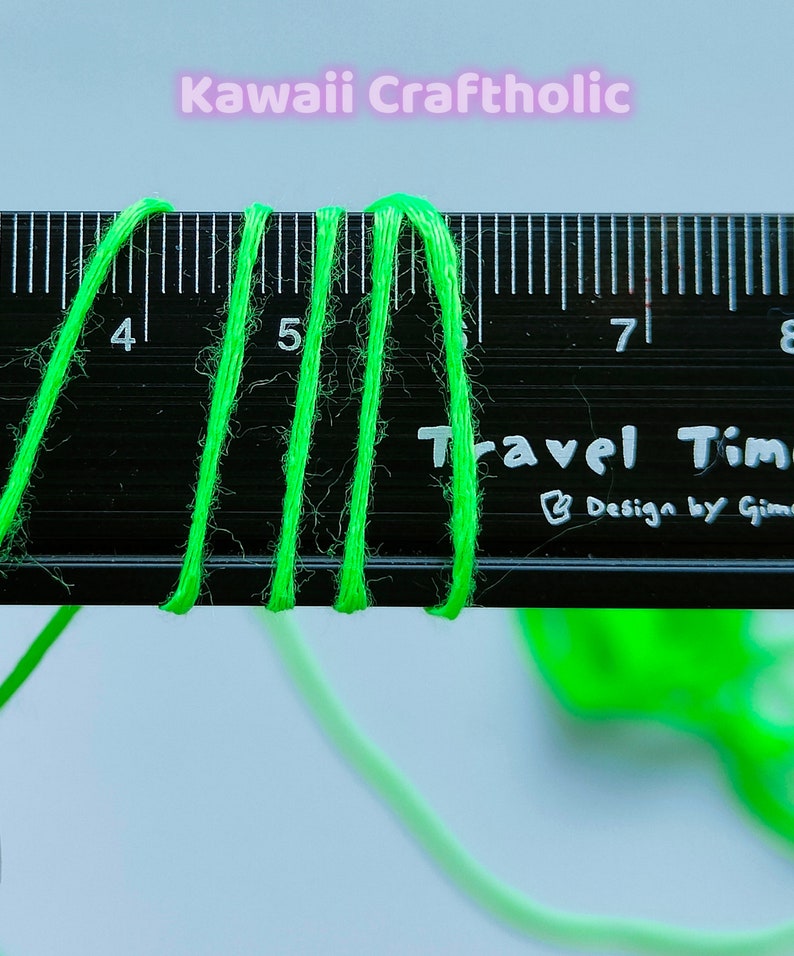 May include: Close-up of three strands of bright green yarn measured against a black ruler. The ruler has markings for inches and the numbers 4, 5, 7, and 8 are visible. The text "Travel Time" and "Design by Gimme" are printed on the ruler.