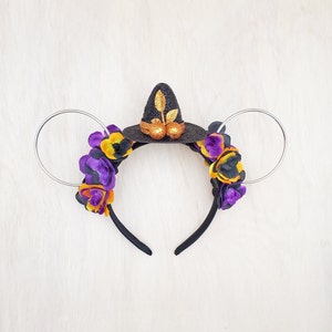 May include: Black witch hat headband with purple and yellow flowers. The headband is made of black felt and has a gold glitter accent. The flowers are made of fabric and are attached to the headband with wire.