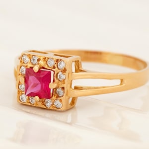May include: A gold ring featuring a square red gemstone set within a halo of smaller clear stones. The band is simple with a double-bar design.