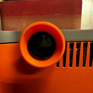 May include: Close-up of an orange dust collection port on a power tool. The port has a black, round opening with a black plastic ring around it.