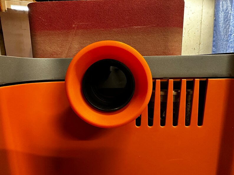 May include: Close-up of an orange dust collection port on a power tool. The port has a black, round opening with a black plastic ring around it.