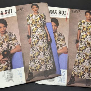 Puede incluir: Múltiples sobres de patrones Vogue Anna Sui con un diseño de vestido con estampado floral. Los sobres muestran a una modelo que lleva el vestido, con el número de patrón V1134 visible. El vestido tiene mangas cortas y una falda larga.