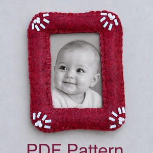 May include: A rectangular dark red felt picture frame featuring hand-stitched edges and white bead accents at each corner. The frame holds a black and white photograph of a smiling infant. The text PDF Pattern is printed below the frame.