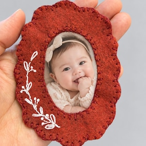 Puede incluir: Un marco ovalado de fieltro color óxido con bordes festoneados y costuras de festón en rojo oscuro. El marco presenta ramas de flores bordadas en blanco en el lado izquierdo y contiene la fotografía de un bebé sonriente con una diadema crema.