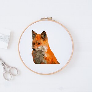 May include: A close-up of a fox cross-stitch embroidery pattern in a wooden embroidery hoop. The fox is orange and brown with a white background.