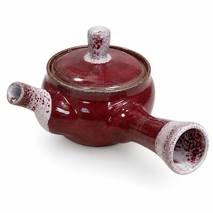 Puede incluir: Una tetera de cer&aacute;mica con asa lateral, con un esmalte rojo brillante y un acabado moteado blanco en contraste en el pomo de la tapa, el pico y el extremo del asa. La tetera tiene un cuerpo redondeado y una tapa circular a juego.