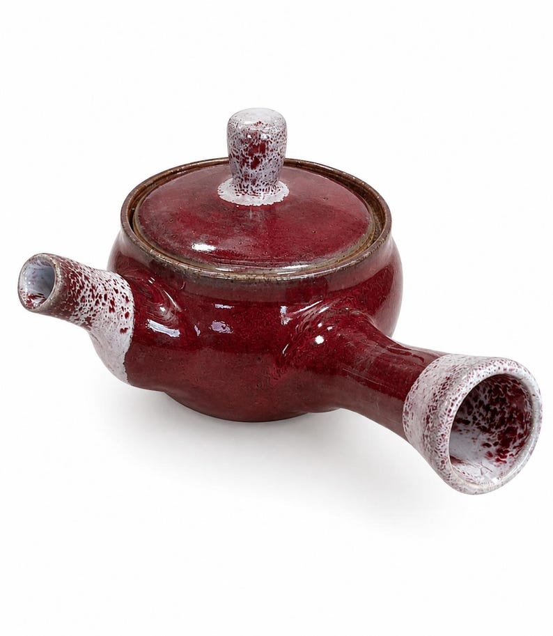 Puede incluir: Una tetera de cer&aacute;mica con asa lateral, con un esmalte rojo brillante y un acabado moteado blanco en contraste en el pomo de la tapa, el pico y el extremo del asa. La tetera tiene un cuerpo redondeado y una tapa circular a juego.