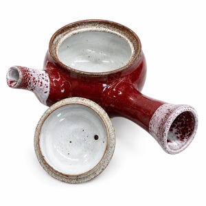 Puede incluir: Una tetera de cer&aacute;mica con asa lateral, con un esmalte rojo intenso brillante en el exterior y un interior blanco moteado. La tapa est&aacute; separada, mostrando una parte inferior blanca con un peque&ntilde;o orificio. El pico y el asa tienen detalles blancos.