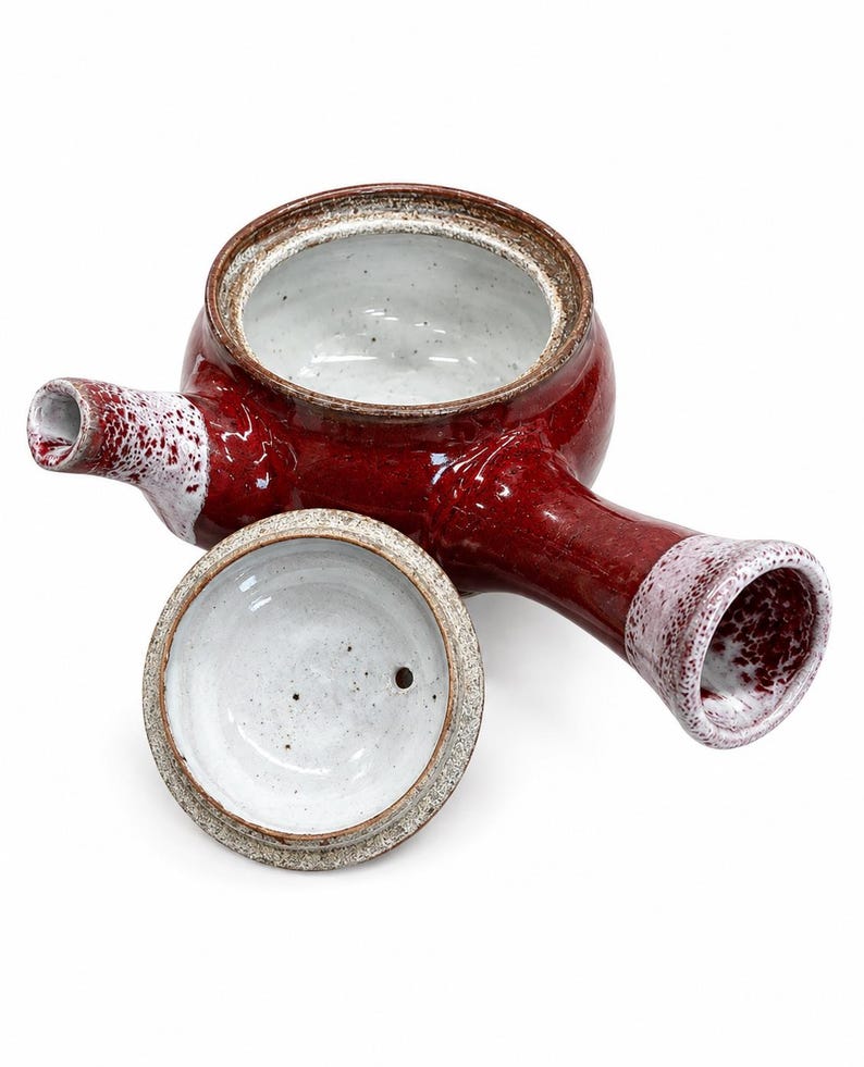 Puede incluir: Una tetera de cer&aacute;mica con asa lateral, con un esmalte rojo intenso brillante en el exterior y un interior blanco moteado. La tapa est&aacute; separada, mostrando una parte inferior blanca con un peque&ntilde;o orificio. El pico y el asa tienen detalles blancos.