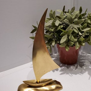 May include: A gold-colored metal sailboat sculpture with a sail and a base that resembles a wave. The sculpture is on a white surface.