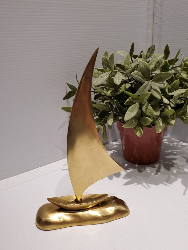 May include: A gold-colored metal sailboat sculpture with a sail and a base that resembles a wave. The sculpture is on a white surface.