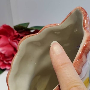 May include: A white ceramic planter shaped like a flower with pink, red, and yellow painted details. The planter has a scalloped rim and a smooth, white interior.